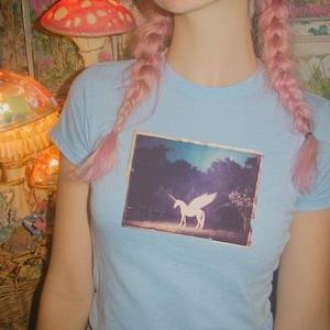 May include: A light blue short-sleeved t-shirt featuring a rectangular graphic print on the chest. The print depicts a white winged unicorn standing in a dark, wooded landscape under a bright sky. The shirt has a crew neckline and a fitted cut.