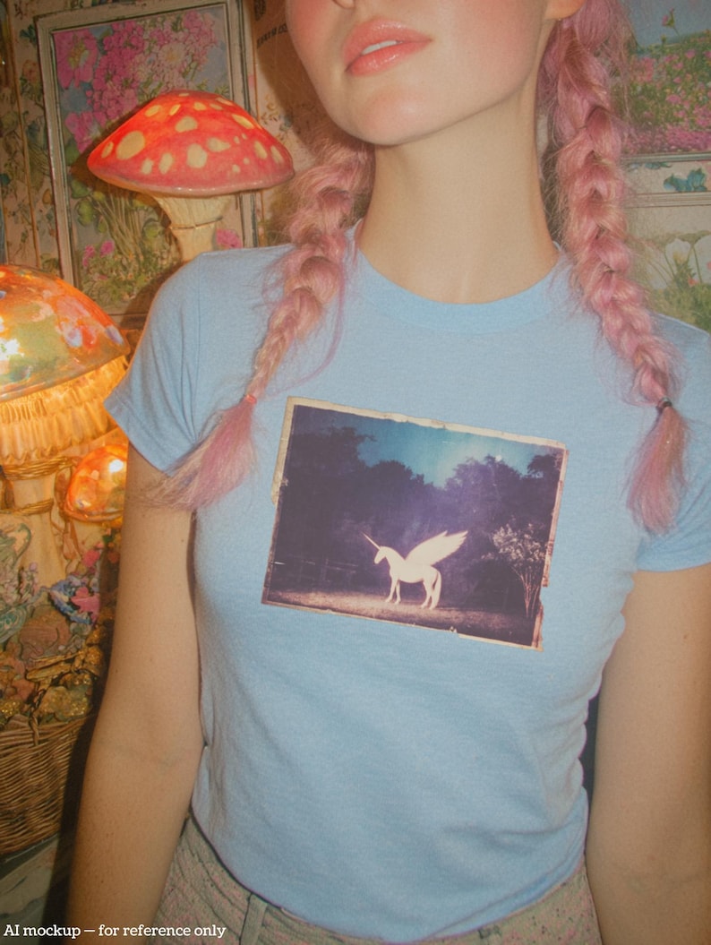 May include: A light blue short-sleeved t-shirt featuring a rectangular graphic print on the chest. The print depicts a white winged unicorn standing in a dark, wooded landscape under a bright sky. The shirt has a crew neckline and a fitted cut.