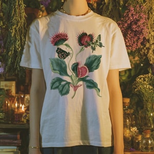 May include: A white short-sleeved t-shirt featuring a central graphic print of a botanical illustration. The design displays green leaves, pinkish-red flowering plants, and two butterflies with detailed wing patterns in shades of black and green.