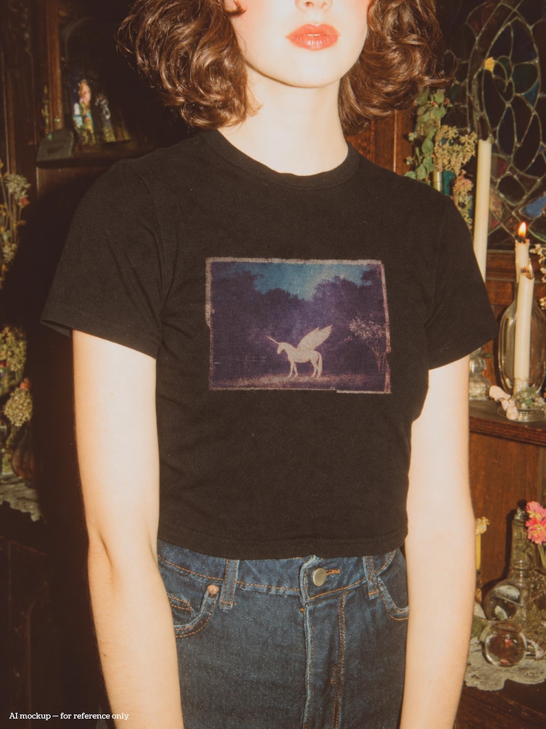 May include: A black short-sleeved t-shirt featuring a rectangular graphic print on the chest. The print depicts a white winged unicorn standing in a dark, wooded landscape under a night sky. The shirt is paired with dark blue denim jeans.