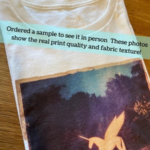May include: A white t-shirt featuring a rectangular graphic print of a winged unicorn in a dark landscape. The shirt has a crew neckline with the text Hazel and Vesper printed on the inside collar. The graphic shows a light-colored unicorn.