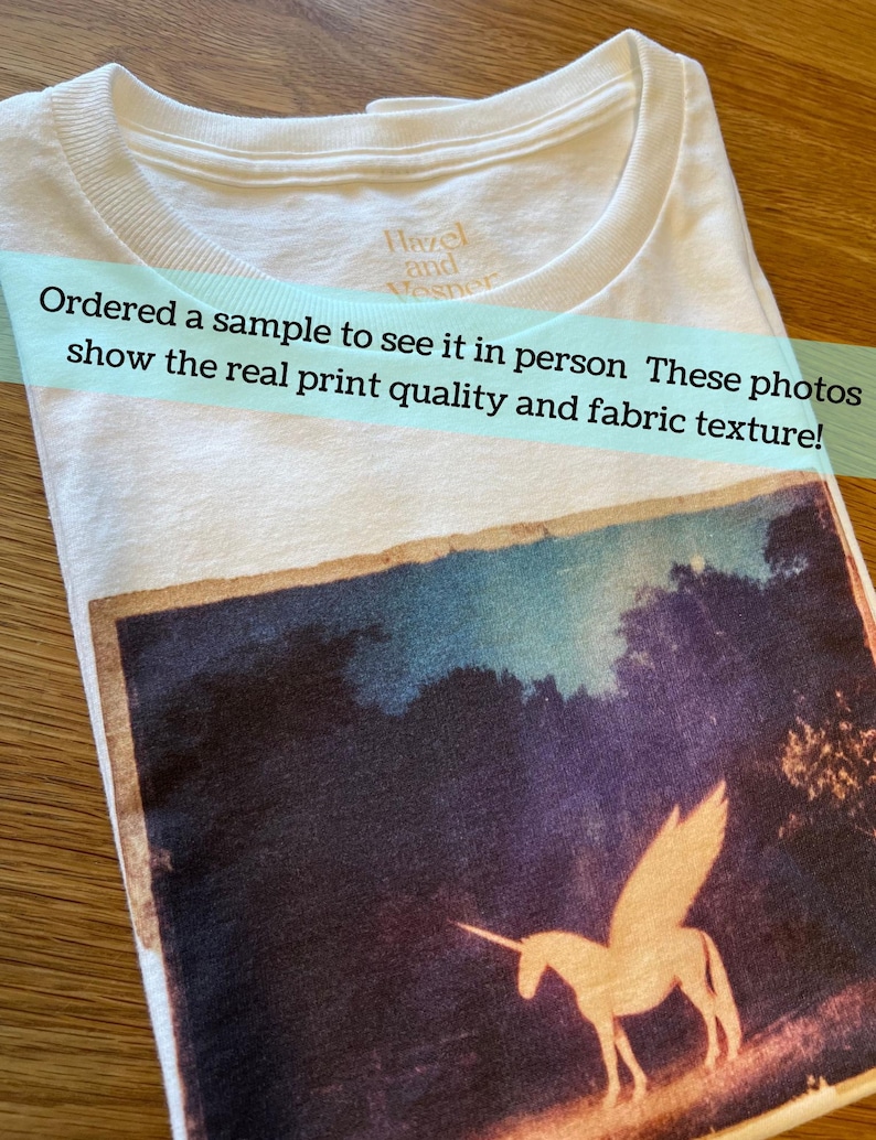 May include: A white t-shirt featuring a rectangular graphic print of a winged unicorn in a dark landscape. The shirt has a crew neckline with the text Hazel and Vesper printed on the inside collar. The graphic shows a light-colored unicorn.