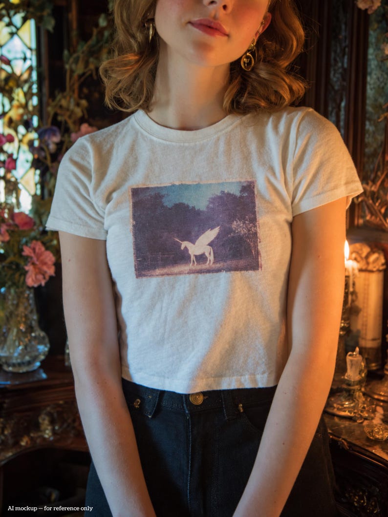 May include: A white short-sleeved t-shirt featuring a rectangular graphic print on the chest. The print depicts a white winged unicorn standing in a dark, wooded landscape with a blue and purple sky. The shirt has a crew neckline and a cropped hem.