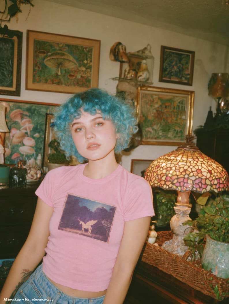 May include: A light pink short-sleeved t-shirt featuring a rectangular graphic print of a winged unicorn standing in a dark forest setting. The shirt has a crew neckline and a fitted silhouette, constructed from a solid-colored cotton fabric.
