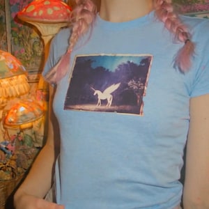 May include: A light blue short-sleeved t-shirt featuring a rectangular graphic print on the chest. The print depicts a white winged unicorn standing in a dark, wooded landscape under a blue sky. The shirt has a crew neckline and a fitted silhouette.