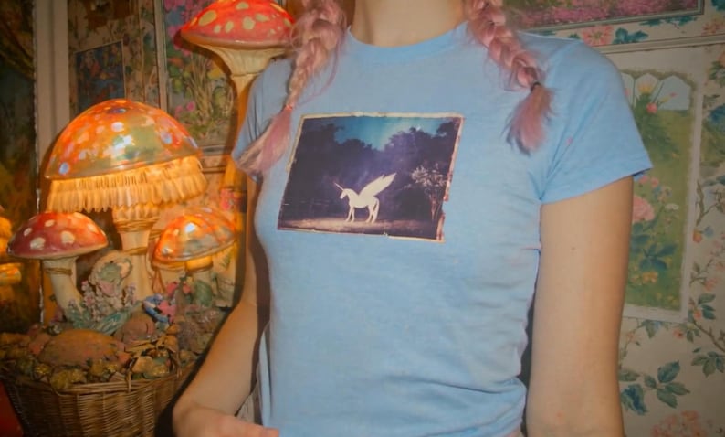 May include: A light blue short-sleeved t-shirt featuring a rectangular graphic print on the chest. The print depicts a white winged unicorn standing in a dark, wooded landscape under a blue sky. The shirt has a crew neckline and a fitted silhouette.