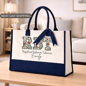 May include: A canvas tote bag with navy blue accents and handles. The bag features the letters "RBT" filled with a floral pattern, along with the text "Registered Behavior Technician" and a name. A navy blue ribbon is tied to the handles.