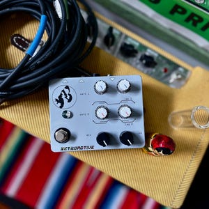 May include: A light grey RETROACTIVE guitar effects pedal with several knobs and a footswitch. The pedal features a black and white graphic. A black cable with a blue band, a clear glass tube, and a red and gold connector are also visible.