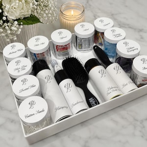 Puede incluir: Una bandeja rectangular blanca que contiene varios artículos de cuidado personal en frascos transparentes con tapas blancas etiquetadas con texto en cursiva negra, incluyendo laca, desodorante, un cepillo negro, quitapelusas y varios artículos.