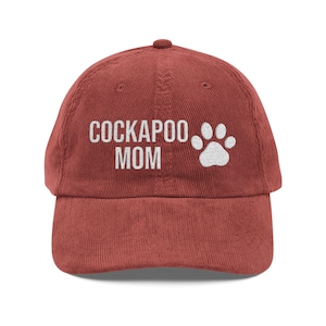 May include: A brick-red corduroy baseball cap with the words "COCKAPOO MOM" embroidered in white, along with a white paw print. The cap has a curved brim and a fabric-covered adjustable strap.