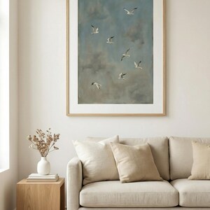 May include: A framed artwork depicts seagulls in flight against a cloudy blue sky. The painting is displayed above a beige sofa with matching pillows. A wooden side table holds a white vase with dried flowers.