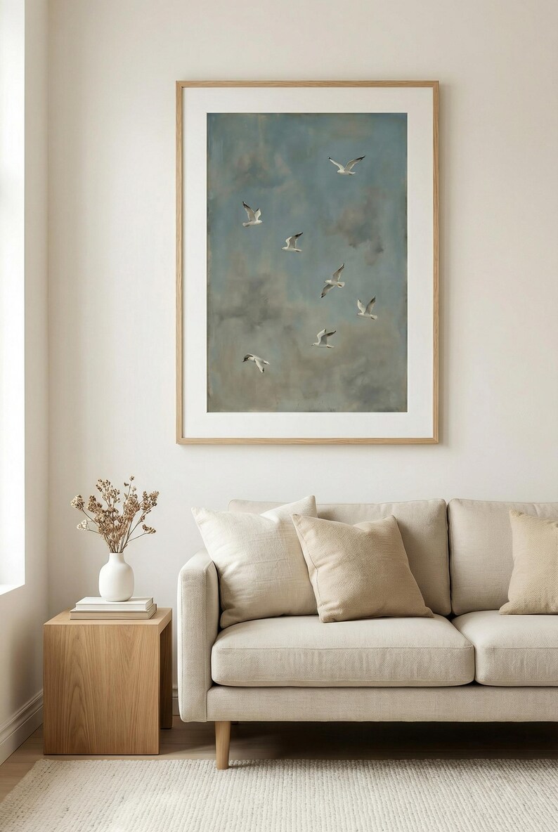 May include: A framed artwork depicts seagulls in flight against a cloudy blue sky. The painting is displayed above a beige sofa with matching pillows. A wooden side table holds a white vase with dried flowers.