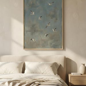 May include: A framed artwork featuring a flock of seagulls soaring against a cloudy, blue sky. The painting has a muted color palette with a wooden frame. The artwork is displayed above a bed with cream-colored bedding and a wooden nightstand.