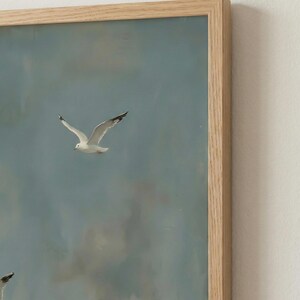 May include: A framed painting depicts seagulls in flight against a cloudy, blue sky. The artwork features several white birds with black wingtips, captured mid-flight. The frame is made of light-colored wood.