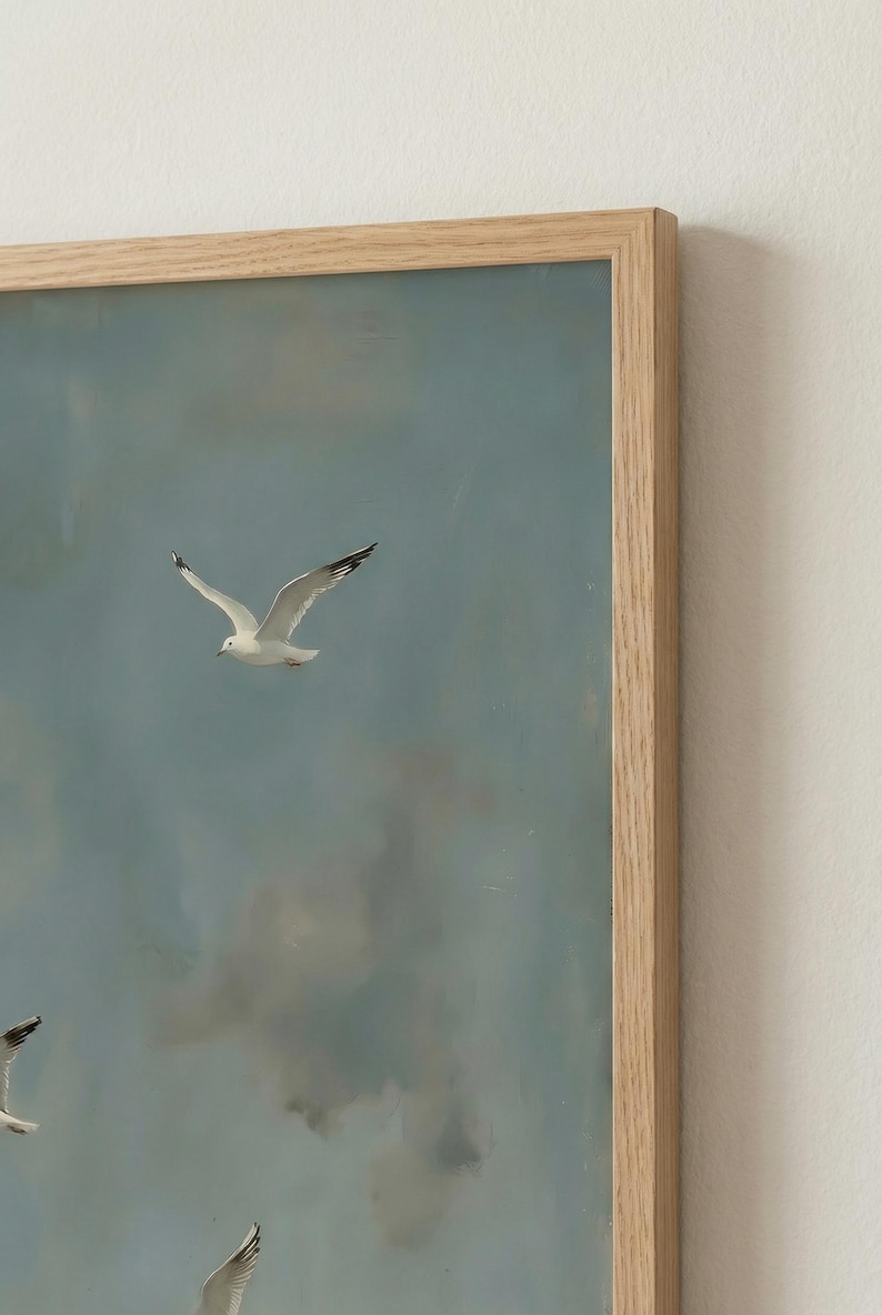May include: A framed painting depicts seagulls in flight against a cloudy, blue sky. The artwork features several white birds with black wingtips, captured mid-flight. The frame is made of light-colored wood.