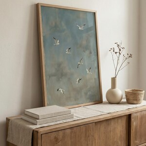 May include: A framed artwork featuring a blue sky with white clouds and several seagulls in flight. The artwork is displayed on a wooden cabinet with a beige vase, a small woven basket, and a stack of books.