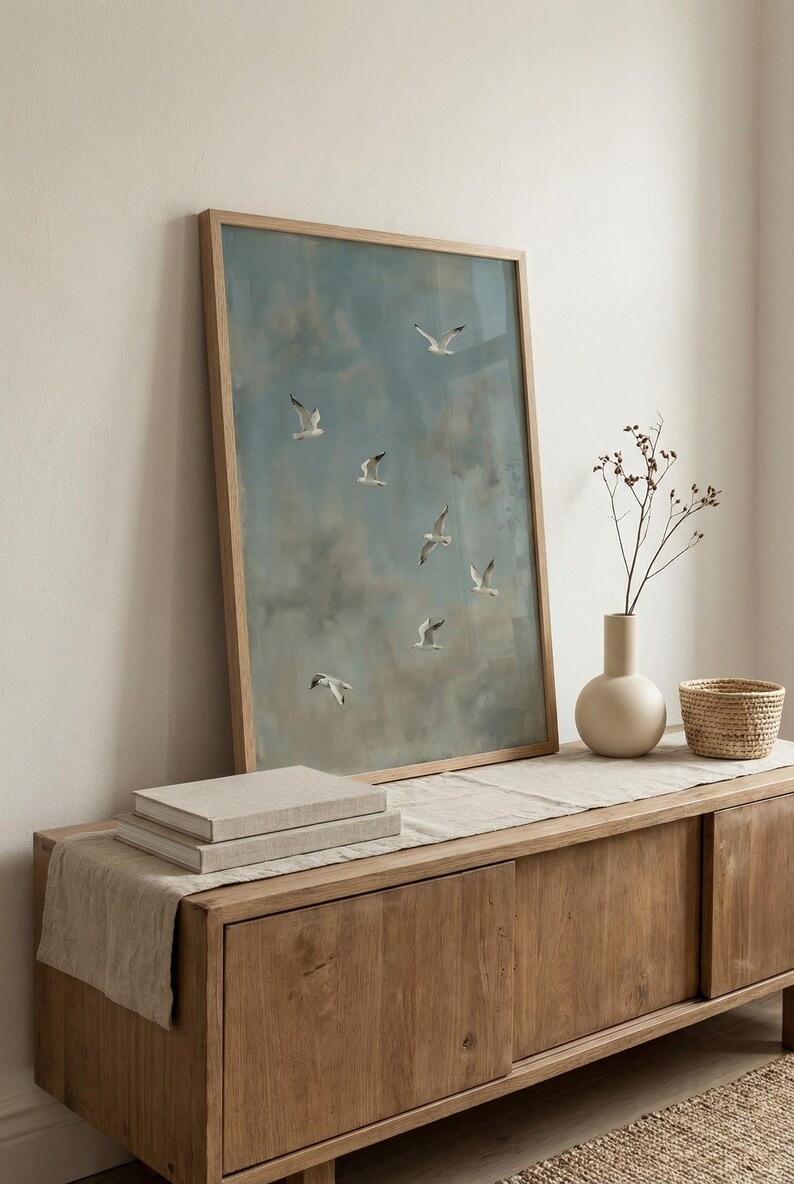 May include: A framed artwork featuring a blue sky with white clouds and several seagulls in flight. The artwork is displayed on a wooden cabinet with a beige vase, a small woven basket, and a stack of books.