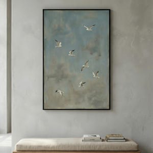 May include: A framed painting depicts several white seagulls in flight against a cloudy blue sky. The artwork is displayed above a wooden bench with a beige cushion. Books are placed on the bench.