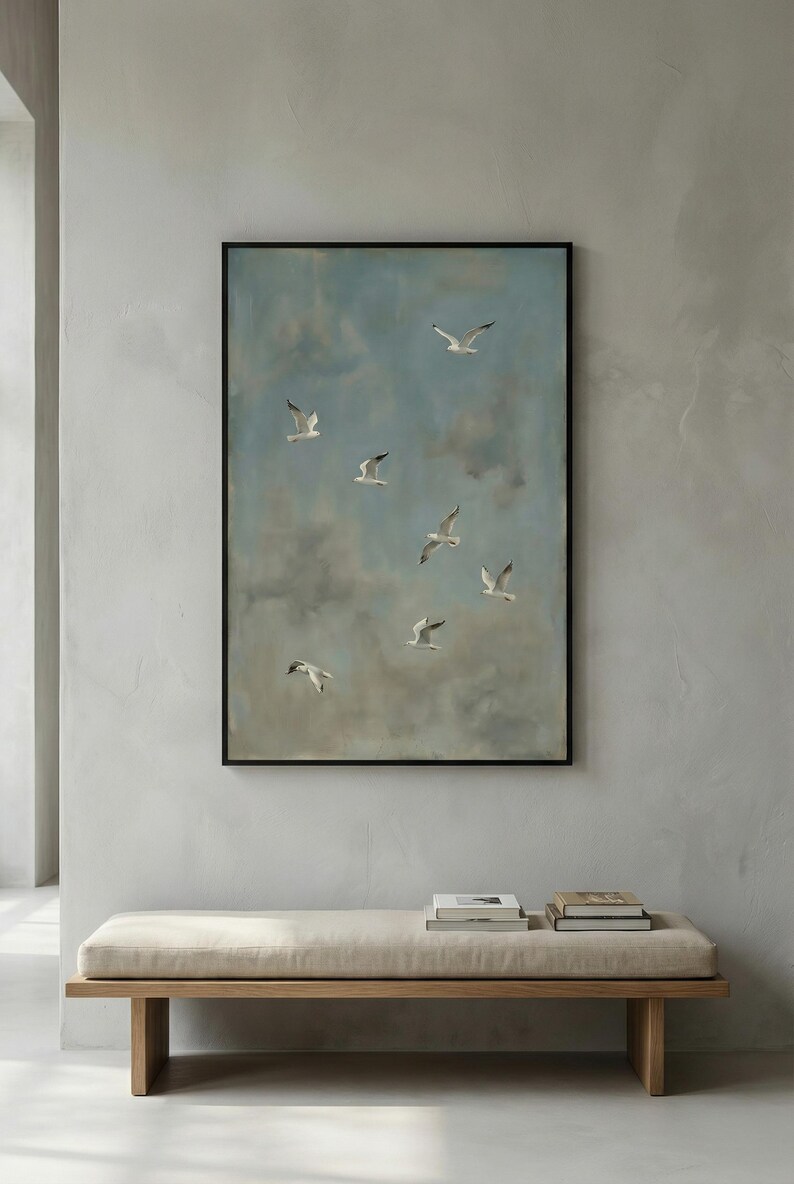 May include: A framed painting depicts several white seagulls in flight against a cloudy blue sky. The artwork is displayed above a wooden bench with a beige cushion. Books are placed on the bench.