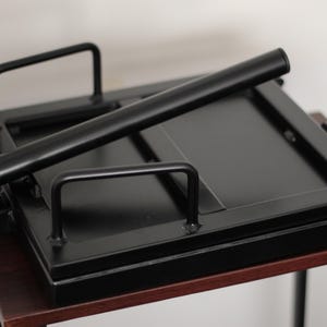 May include: A black metal press with a rectangular base and a hinged top. It features a long, cylindrical handle and two smaller handles. The press is set on a dark wooden table.