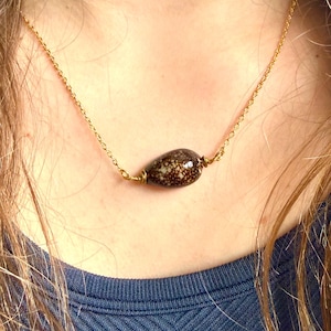 May include: A gold chain necklace with a teardrop-shaped pendant. The pendant is brown with a speckled pattern and is suspended from the chain. The necklace is worn on a person wearing a blue shirt.