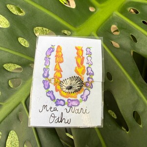 May include: A handmade necklace featuring a seashell pendant, gold chain, and colorful floral accents. The necklace is displayed on a card with the text "Mea Wari Oahu" and is set against a large green leaf.
