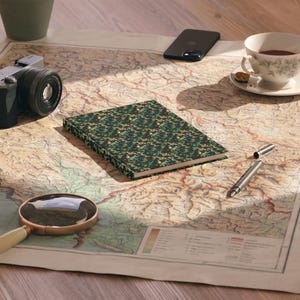 May include: A patterned journal with a camouflage design in shades of green, black, and gold rests on a vintage map. A camera, a magnifying glass, a pen, a smartphone, and a teacup are also on the map.