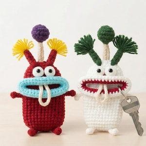 以下が含まれることがあります： 手作りの編み物モンスターキーホルダー2つ。1つは青い口と黄色のフリンジが付いた赤色、もう1つは赤い口と緑色のフリンジが付いた白色です。どちらも口の中に鍵が入っています。