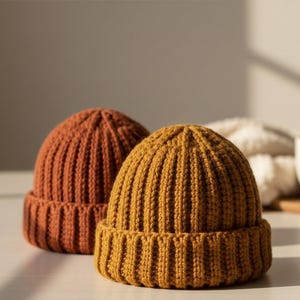 Puede incluir: Dos gorros de punto acanalado. Uno es de un color naranja cálido y terroso, y el otro es amarillo dorado. Los sombreros están hechos de un hilo grueso y texturizado y tienen un borde doblado. Los gorros se exhiben sobre una superficie clara.