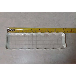 May include: A clear, rectangular acrylic stamp block with a wavy edge, featuring a grid pattern. A yellow tape measure is placed next to the block, showing measurements in inches.