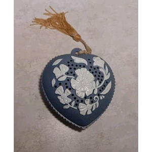 Puede incluir: Infusor de té azul en forma de corazón con detalles florales blancos en relieve. El infusor tiene pequeños agujeros para infusionar el té y está rematado con una borla beige. El diseño recuerda a la porcelana clásica.