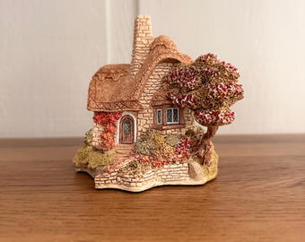 May include: A miniature stone cottage figurine featuring a textured thatched roof, a central chimney, and a brown wooden door. The exterior is detailed with a small bay window, climbing vines, and a sculpted tree with red and brown foliage.