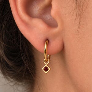 May include: A gold hoop earring featuring a small, dangling charm in a diamond shape. The charm is framed in gold and contains a single, round, deep red gemstone set in the center. The hoop is polished and smooth, creating a reflective surface.