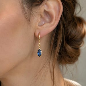 May include: A gold hoop earring featuring a marquise-cut blue gemstone pendant. The earring consists of a thin circular gold hoop that connects to a small, faceted, deep blue stone encased in a gold bezel setting, hanging vertically from the hoop.