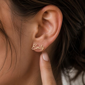 May include: A gold-toned stud earring featuring the name Emily written in a cursive script font. The earring is positioned on the earlobe, showcasing the polished metallic finish and the flowing, connected lettering of the custom name design.