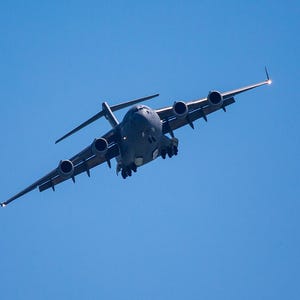 May include: A large, gray military transport aircraft, seen from below, is in flight against a clear, bright blue sky. The plane has four engines and a high wing design. The image is taken from a low angle, emphasizing the size of the aircraft.