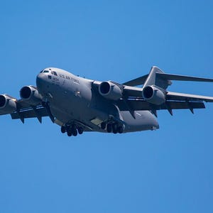 May include: A large gray U.S. Air Force C-17 Globemaster III military transport aircraft in flight against a clear blue sky. The aircraft has four engines and a high-wing design, with the text "U.S. AIR FORCE" visible on the fuselage.