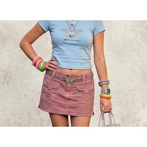 May include: A light blue t-shirt with a star design, worn with a pink denim mini skirt. The outfit is accessorized with a butterfly belt, colourful bracelets, and a watch. A clear tote bag is held in the hand.
