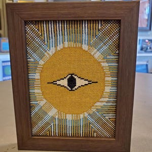 Puede incluir: Obra de arte en punto de cruz enmarcada. Presenta un sol amarillo con un diseño de ojo en blanco y negro. El sol está rodeado de líneas radiantes en amarillo, azul y blanco. El marco es de madera marrón oscuro.