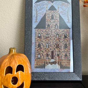Puede incluir: Arte de punto de cruz enmarcado de una casa embrujada con un murciélago y árboles desnudos. La casa es de ladrillo con varios personajes espeluznantes en las ventanas. Una linterna de cerámica con forma de calabaza está frente al marco.