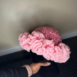 May include: A pink and rose-colored crochet hat with a wide, ruffled brim. The hat is displayed on a black mannequin head. The hat's crown is a mix of pink and rose hues, while the brim is a lighter, ruffled pink.