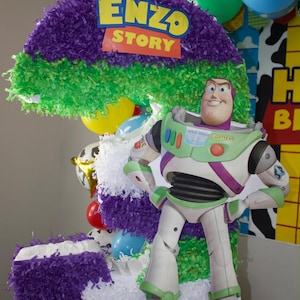 Puede incluir: Una piñata grande con forma de número tres decorada con flecos de papel morado, verde y blanco. La piñata presenta un recorte del personaje Buzz Lightyear y un letrero que dice ENZO STORY en letras amarillas y rojas sobre un fondo azul.