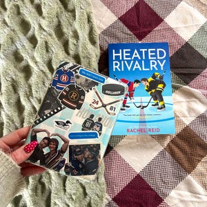 May include: A book titled "Heated Rivalry" with a hockey theme, alongside a phone case decorated with hockey stickers and photos. The book cover shows two hockey players. The phone case has the text "SHANE" and "HOLLANOV".