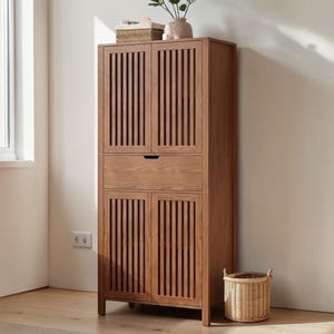 May include: A tall, wooden storage cabinet with a light brown finish. The cabinet has two doors on the top and bottom with vertical slat detailing, a drawer in the middle, and a woven basket on the floor. A small vase and basket sit on top.