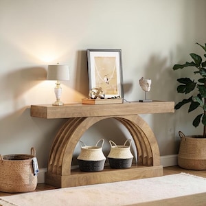 May include: A light brown wooden console table with an arched base, featuring two woven baskets with black and white details. The table is decorated with a lamp, framed artwork, and decorative objects. A potted plant and a woven basket are on the floor.