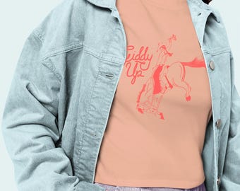 May include: A peach-colored t-shirt with a red illustration of a rodeo rider on a bucking bronco, with the words "Riddy Up" in red script. The model is wearing a light blue corduroy jacket and purple jeans.