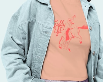 May include: A peach-colored t-shirt with a red illustration of a rodeo rider on a bucking bronco, with the words "Riddy Up" in red script. The model is wearing a light blue corduroy jacket and purple jeans.