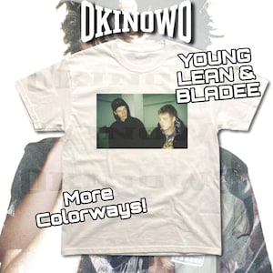 May include: A white t-shirt featuring a rectangular image of two people. The text "OKINOWO" is at the top, with "YOUNG LEAN & BLADEE" on the right side. Below the image is the text "More Colorways!"
