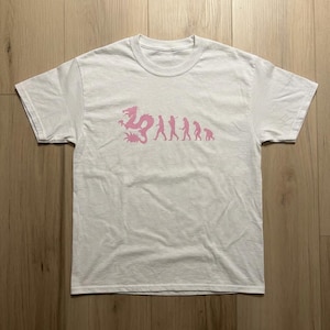 Peut inclure: T-shirt blanc avec un motif rose représentant un dragon se transformant en silhouettes humaines, symbolisant l'évolution. Le t-shirt a un col rond et est fait d'un tissu doux.