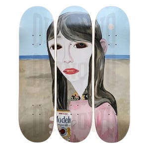 May include: A triptych of three skateboard decks featuring a continuous illustration of a person with long dark hair, wearing a pink shirt with a patterned collar, holding a can of Modelo Especial beer against a tan beach and blue sky background.